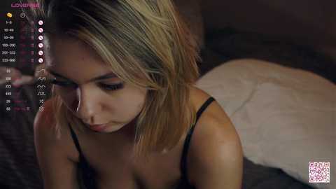 Media: A video of a blonde woman with medium skin tone, wearing a black bra, lying on a bed with a white pillow, in a dimly lit room.