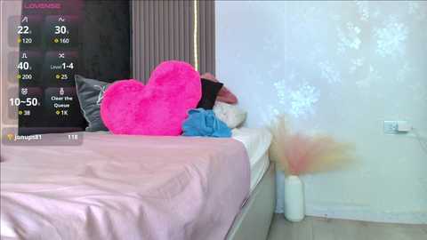 Media: Video of a tidy, pastel-colored bedroom with a bed featuring a large pink heart pillow, a stuffed animal, and a fluffy white pillow. The background has a white wall and a digital display showing weather and temperature data.