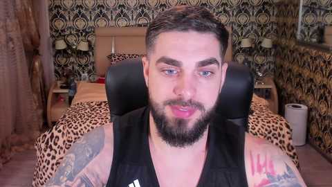 Media: Video of a bearded, tattooed man with short dark hair, wearing a black tank top, sitting on a leopard-print bed in a room with patterned wallpaper.