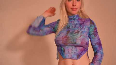 Media: Video of a blonde woman with fair skin, wearing a long-sleeved, tie-dye crop top in shades of blue, purple, and pink, standing against a beige wall.