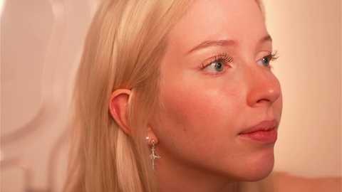 Media: Video of a young Caucasian woman with long, straight blonde hair, fair skin, and blue eyes. She has a neutral expression, wearing small, delicate silver earrings. Background is blurred, beige, and softly lit.