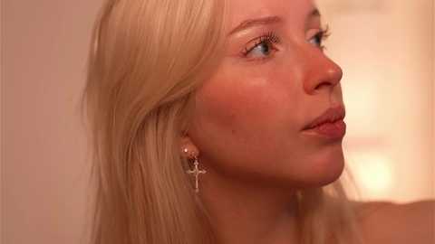 Media: Video of a young, fair-skinned, blonde woman with blue eyes, wearing a delicate gold cross earring. She has smooth skin and is seen from the side, looking slightly upward. The background is soft and blurred.