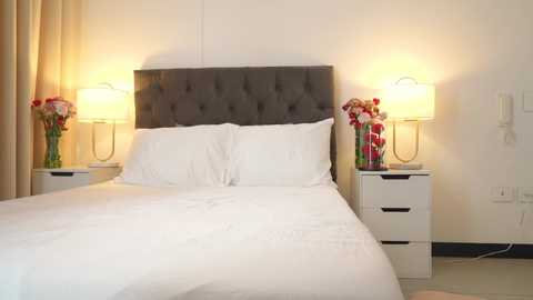 Media: A video of a modern, minimalist bedroom with a tufted gray headboard, white linens, and two white bedside tables each holding a yellow lamp and a bouquet of red and white flowers. The background is a plain, off-white wall.