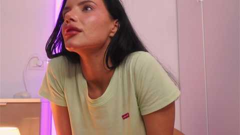 Media: Video of a young woman with long black hair, fair skin, wearing a light green t-shirt, looking contemplative in a minimalist room with purple and white lighting.