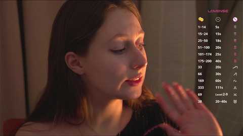 Media: A video of a young woman with fair skin and straight brown hair, wearing a black top, smiling softly. The background is blurred, and a digital clock displays the time and date.