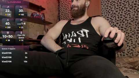 Video of a muscular, bearded man in a black tank top with white text, sitting on a couch, holding a controller, surrounded by a virtual reality setup.