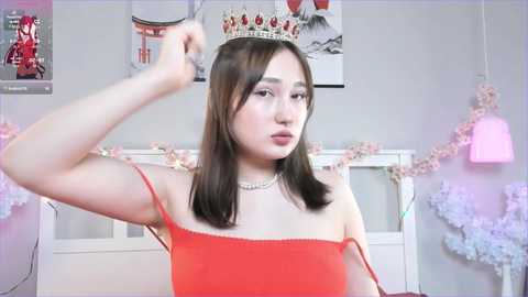 Media: Video of an East Asian woman with straight, shoulder-length brown hair, wearing a red strapless dress, adjusting a tiara in a pastel-colored room adorned with floral decorations and pink lights.