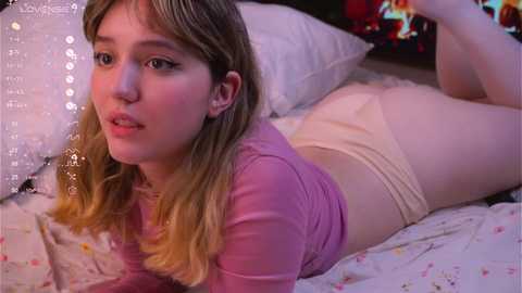 Media: A video of a young Caucasian woman with light skin and long blonde hair, wearing a pink top and beige shorts, lying on a bed with white and floral-patterned sheets, in a cozy, dimly-lit bedroom.