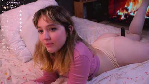 Video of a young woman with light skin, blonde hair, and bangs, lying on a bed in a snug, cozy room. She wears a purple top and white panties, with a TV playing a fire-themed video game in the background.