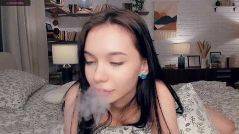 Video of an Asian woman with long black hair, wearing blue earrings, lying on a bed smoking, surrounded by modern decor, white brick wall, and abstract art.