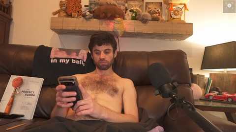 Media: Video of a bearded, shirtless, fair-skinned man with dark hair, sitting on a brown leather couch, reading a book, wearing headphones, surrounded by a cozy living room with wooden decor, a lamp, and a fireplace.