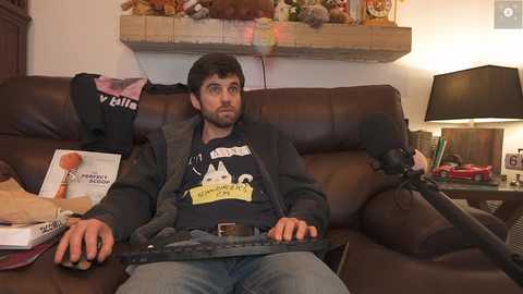 Media: Video of a man with short dark hair and beard, wearing a black t-shirt and gray jacket, sitting on a brown leather couch, holding a keyboard, surrounded by books and a lamp.