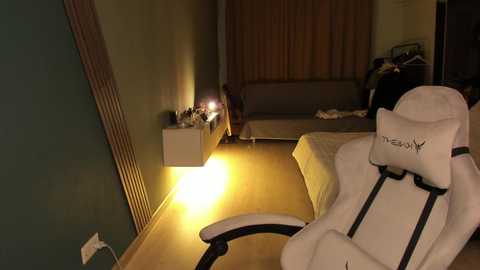 Video of a dimly lit bedroom with a white gaming chair, beige walls, a bed, and a nightstand with a lamp and bottle, wooden floor, and a window with beige curtains.