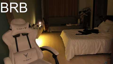 Video of a dimly-lit bedroom with a white gaming chair, a neatly made bed, and a lamp emitting a warm glow.