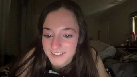 Media: Video of a smiling young woman with fair skin and long brown hair, in a dimly lit bedroom with messy bed and furniture.