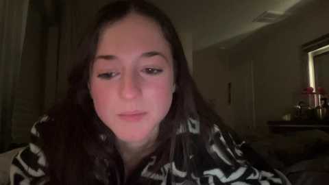 Media: Video of a young woman with long dark hair, wearing a black and white patterned shirt, looking slightly downcast, in a dimly lit bedroom with a bed and a window visible in the background.