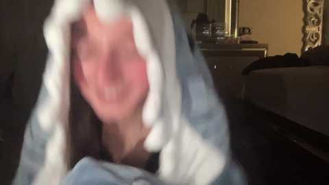 Media: Video of a person in a blue hooded cloak, head covered, with a blurred face, standing indoors near a bed and dresser.