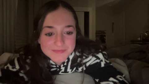 Media: Video of a young woman with long, curly dark hair and fair skin, smiling softly. She wears a black and white patterned sweater. Background shows a dimly lit bedroom with a bed, dresser, and dark curtains.