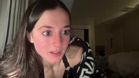 Media: Video of a young woman with long brown hair, light skin, and brown eyes, wearing a black and white striped shirt, looking slightly to the side. Background includes a dimly lit room with beige walls and a door.