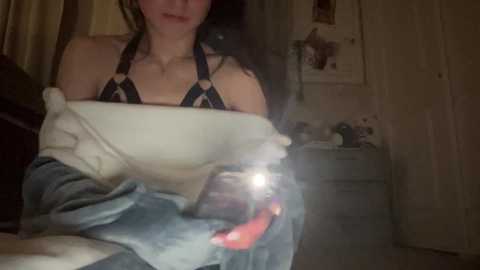 Media: Video of a woman in a dimly lit bedroom, wearing a black and white striped bra, holding a smartphone. She has a slim physique, fair skin, and long dark hair. Background shows a white dresser, a framed poster, and a door.