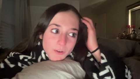 Media: A video of a young woman with long, dark hair, light skin, and wide eyes, looking thoughtful, holding a pillow on a couch. She wears a black and white patterned robe. Background shows a dimly lit room with a doorway and a window.