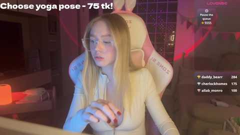 Media: Video of a young, fair-skinned blonde woman with glasses in a white top, seated on a gaming chair. Background includes a dimly lit room with a red glow, gaming console, and chat window with 73.5k viewers.