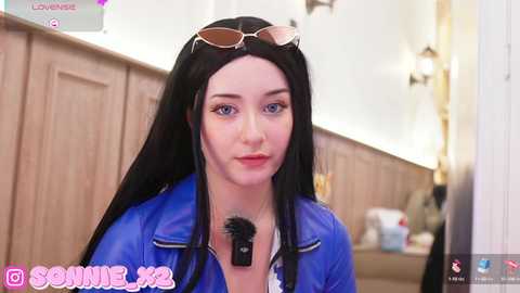 Media: Video of a young woman with long black hair, fair skin, and blue eyes, wearing a blue leather jacket and pink sunglasses. She's indoors, with a wooden wall and a lamp in the background.