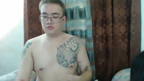 Media: Video of a young, fair-skinned, short-haired Asian man with glasses and tattoos, including a large floral design on his left shoulder, standing shirtless in a room with patterned curtains and bookshelves.