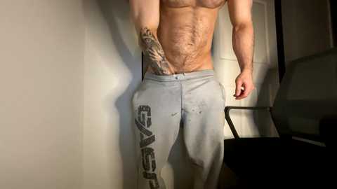 Media: Video of a shirtless man with a muscular physique, wearing grey sweatpants with \"Sweat\" branding on the left leg. He has a tattoo on his right side. Background shows a plain white wall and a partially visible black chair.