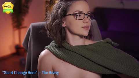 Media: A video of a young, fair-skinned woman with shoulder-length brown hair, wearing glasses and a green sweater, sitting in a chair, with a blurry background. Text reads, \"Short Change Hero - The Heavy.\