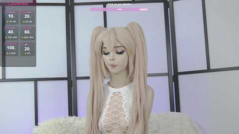 Media: Video of a pale-skinned, petite young woman with long, straight, light-pink hair styled in twin ponytails, wearing a white lace crop top, sitting on a plush, beige cushion. Background includes a black and white room divider with a pink and black screen displaying health bars.