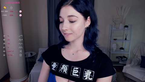 Video of a young woman with pale skin and dark blue hair, wearing a black crop top with white kanji characters, standing indoors near a white bookshelf and a tall plant.