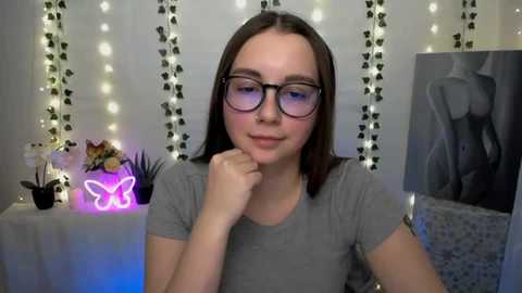 Video of a young woman with light skin and straight brown hair, wearing glasses and a gray t-shirt, standing in a cozy room with fairy lights, a butterfly neon sign, and a framed black-and-white artwork.