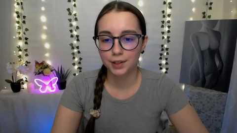 Video of a young woman with fair skin, long brown braid, and glasses, wearing a gray t-shirt, standing in a cozy room with fairy lights, a pink butterfly light, and a black-and-white silhouette painting.