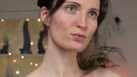 Video of a young woman with fair skin, blue eyes, and dark brown hair styled in an updo. She has a serene expression, with subtle makeup. Background shows a blurred shelf with decorative items and warm lighting.