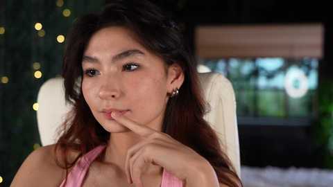 Video of an Asian woman with long dark hair, wearing a pink top, sitting indoors, with blurred greenery and twinkling lights in the background.