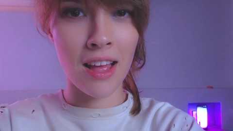 Video of a young, fair-skinned woman with light brown hair, wearing a white shirt, smiling in a dimly lit room with purple and pink lighting.