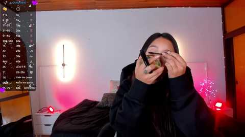 Media: A video of a young Asian woman with long black hair, wearing a black hoodie, hiding her face with her hands, in a dimly lit bedroom with a wooden ceiling, pink and white lights, and a messy bed.