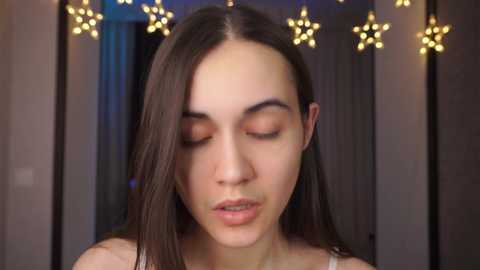 Media: Video of a young woman with straight, long brown hair, closed eyes, and a neutral expression, standing in a dimly lit room with star-shaped lights hanging in the background.