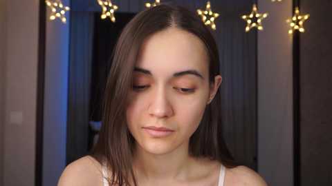 Media: Video of a young woman with fair skin, straight brown hair, wearing a white spaghetti strap top, in a dimly lit room with star-shaped string lights.