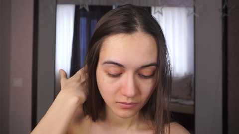 Media: A video of a young woman with light skin and straight, shoulder-length brown hair. She is topless, with her eyes closed and a contemplative expression, as she holds her hair with one hand. The background shows a dimly lit room with a window and curtains.