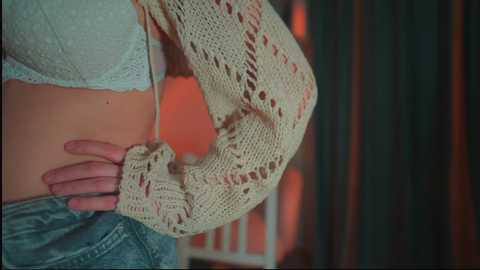 Media: Video of a person wearing a beige crochet cardigan over a white lace bralette and blue jeans, with a hand on their hip, in a dimly lit room with dark curtains and warm lighting.