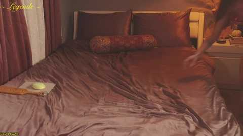 Media: Video of a woman in a plush, dimly lit bedroom, wearing a beige nightgown, adjusting a brown satin bedspread. The room features dark purple curtains, a floral-patterned pillow, and a bedside lamp casting a warm glow.