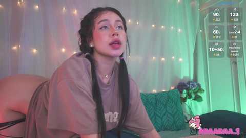 Video of a young Asian woman with long black hair, wearing a brown T-shirt, leaning on a bed with green patterned pillows in a dimly lit room.