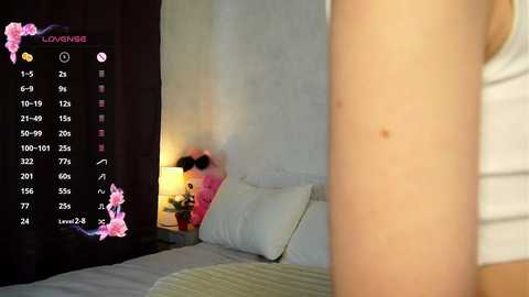 Media: Video of a cozy bedroom with a bed, white pillows, and a pink teddy bear. A close-up of a person's upper body is in the foreground, partially visible. A digital clock with a pink floral frame displays 10:19.