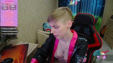 Media: A video of a young, fair-skinned woman with a shaved side and long blonde hair, wearing a black robe with pink trim, sitting at a gaming desk.