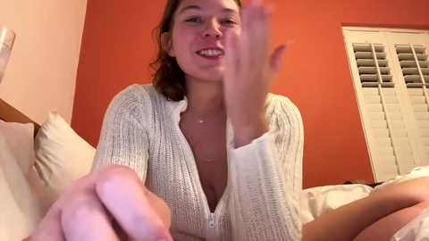 Media: Video of a Caucasian woman with light skin and brown hair, wearing a white knit cardigan, smiling while holding a toy phone in a bedroom with orange walls and white blinds.