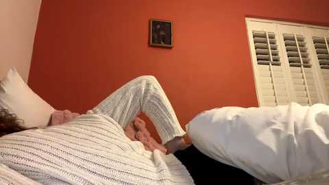 Media: Video of a person in white knitted pajamas lying on a bed with white sheets and a dark headboard against a red wall with a framed picture.