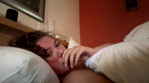 Media: Video of a light-skinned woman with curly brown hair, lying in bed under white sheets, wearing a ring, gazing pensively at a glass of water on a bedside table.