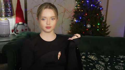 Media: Video of a young Caucasian woman with fair skin and blonde hair, wearing a black sweater, sitting on a green couch. Christmas tree adorned with multicolored lights and ornaments in the background.